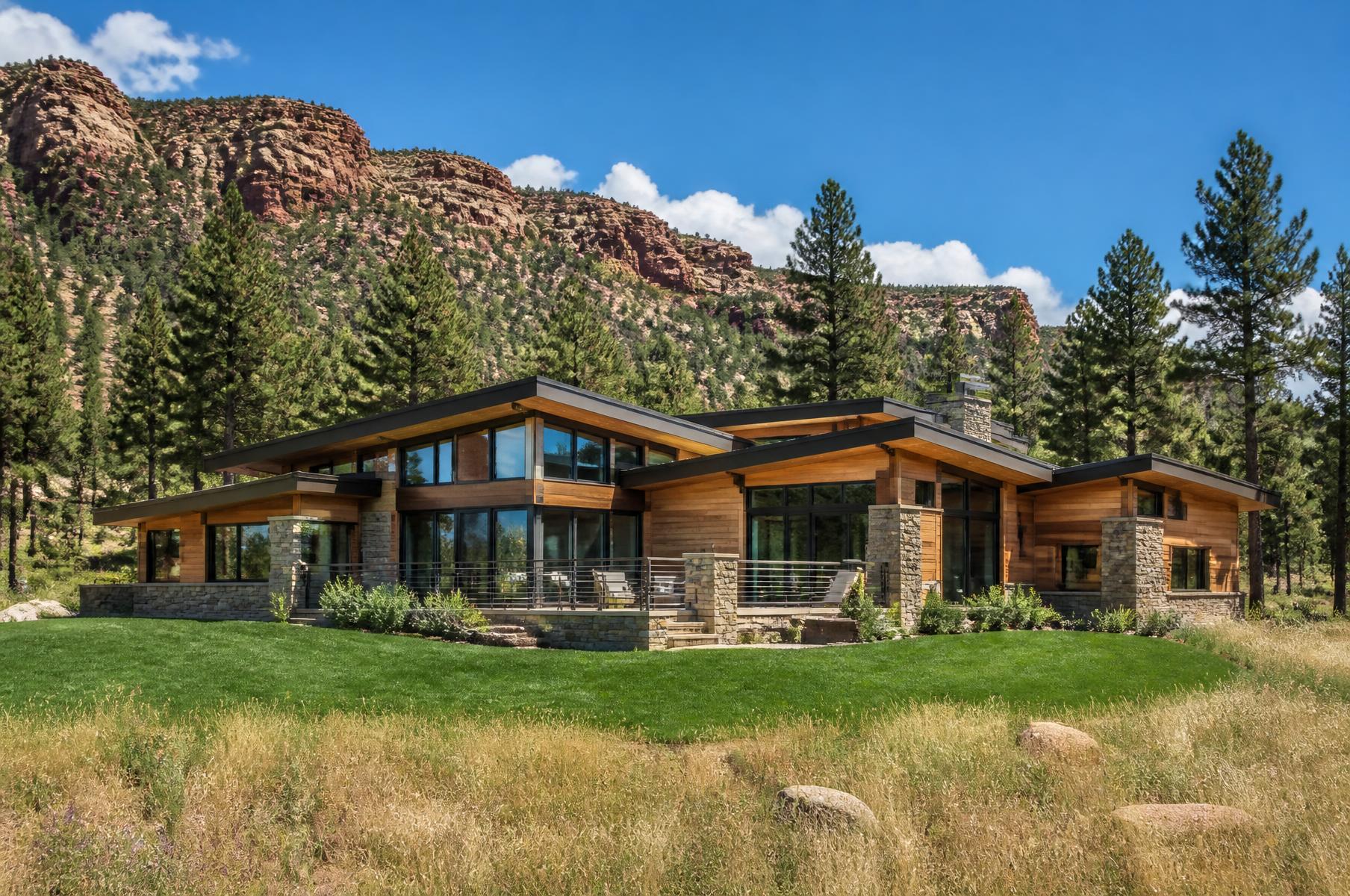 Completed mountain modern home with stone base, cedar cladding and low shed roofs below a red sandstone ridge in summer