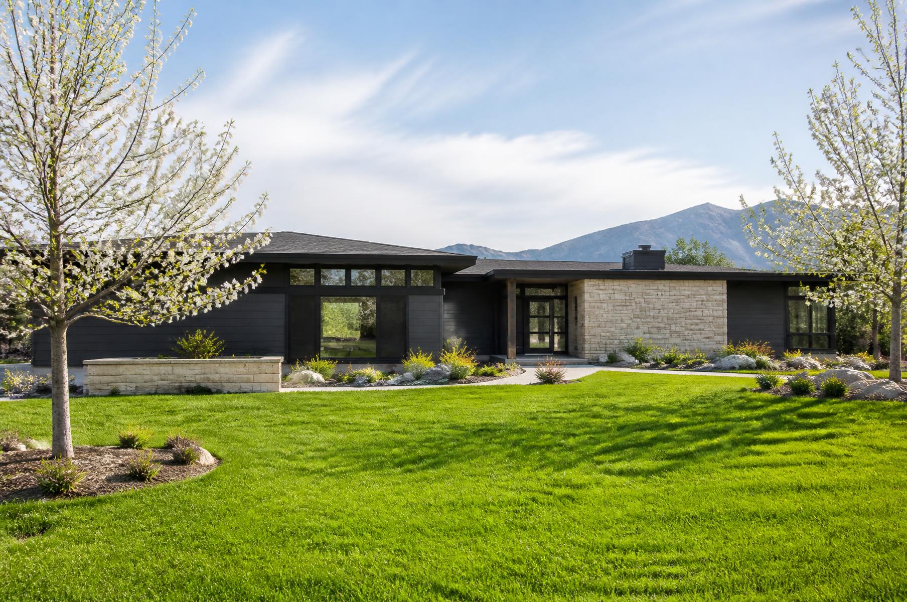 Completed single-story modern ranch home with charcoal siding and a sandstone entry wall behind a bright green spring lawn
