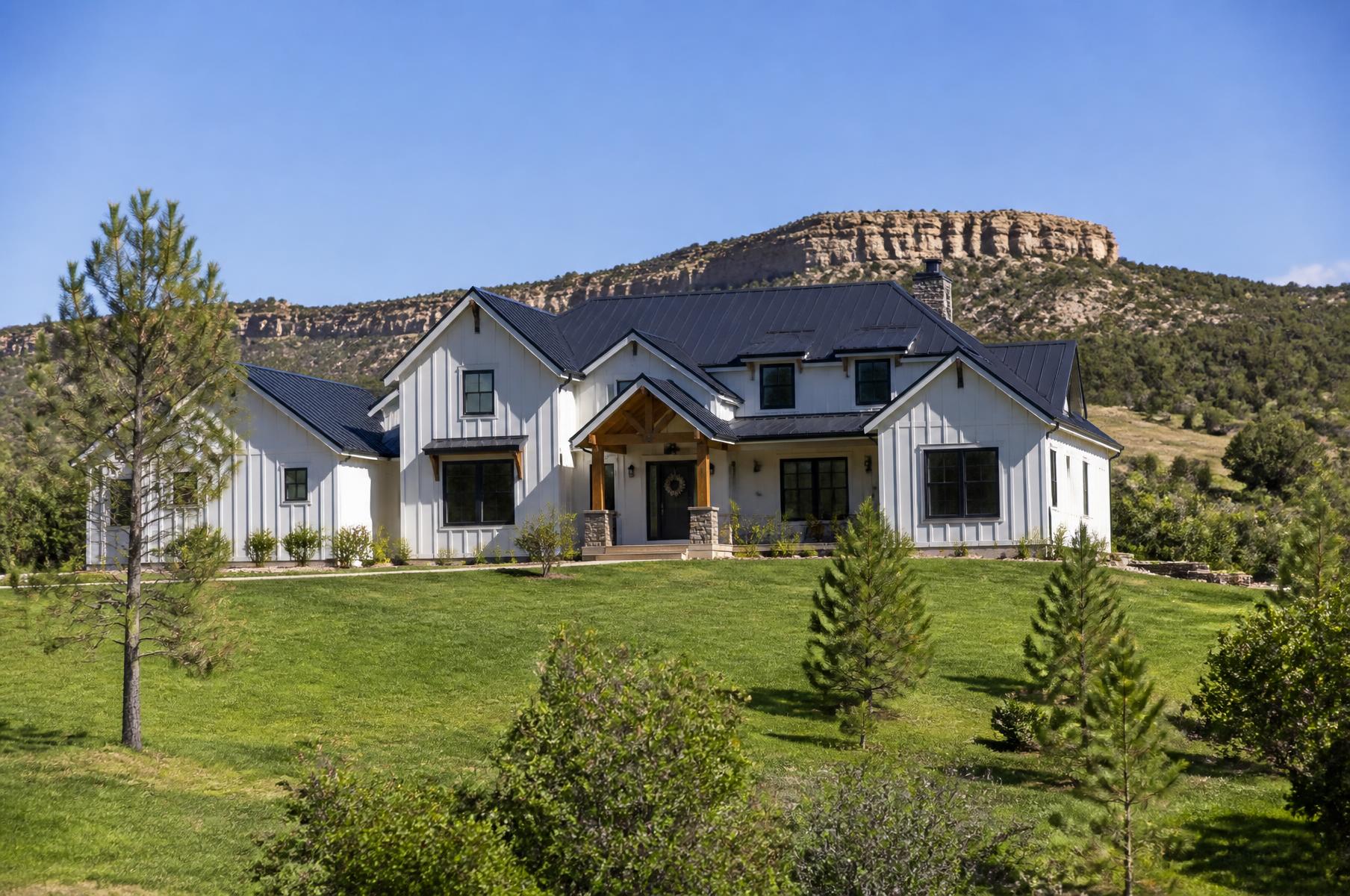 Completed white modern farmhouse with a black metal roof on a green meadow with a sandstone bluff behind in spring