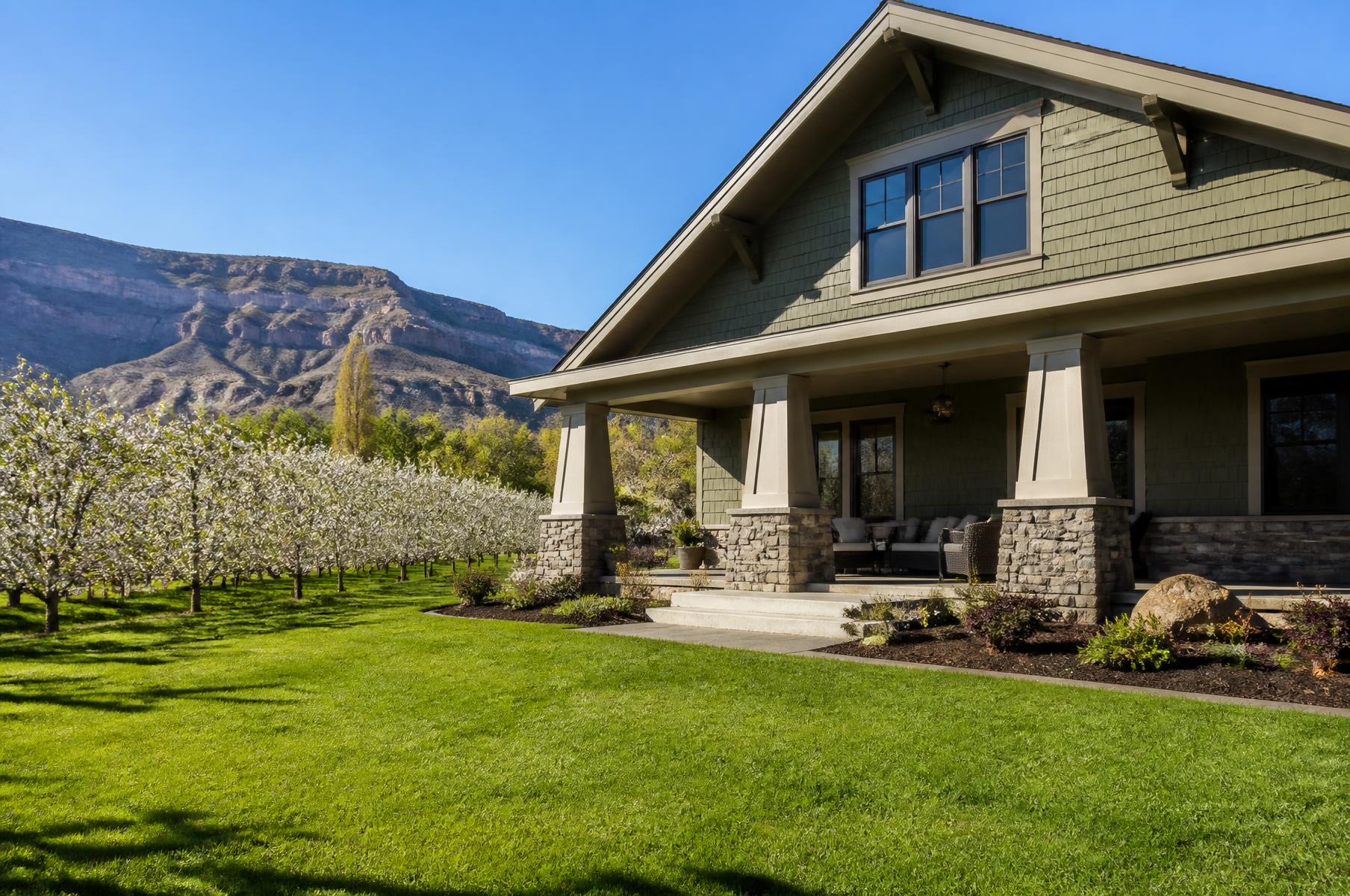 Completed craftsman home with olive shingle siding and a deep porch beside a blossoming orchard in spring