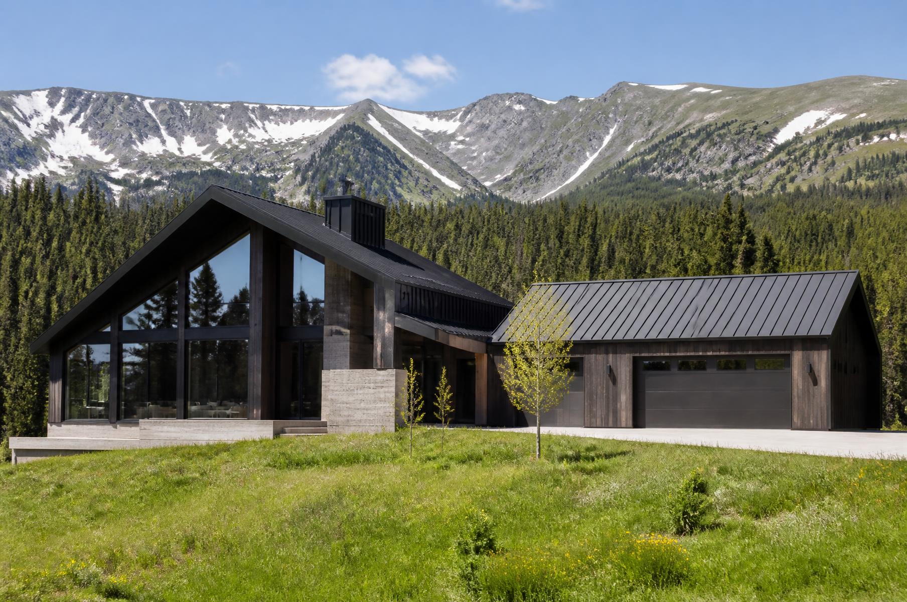 Completed alpine contemporary home with an asymmetric steep roof and timber garage wing on a green meadow below snowy peaks