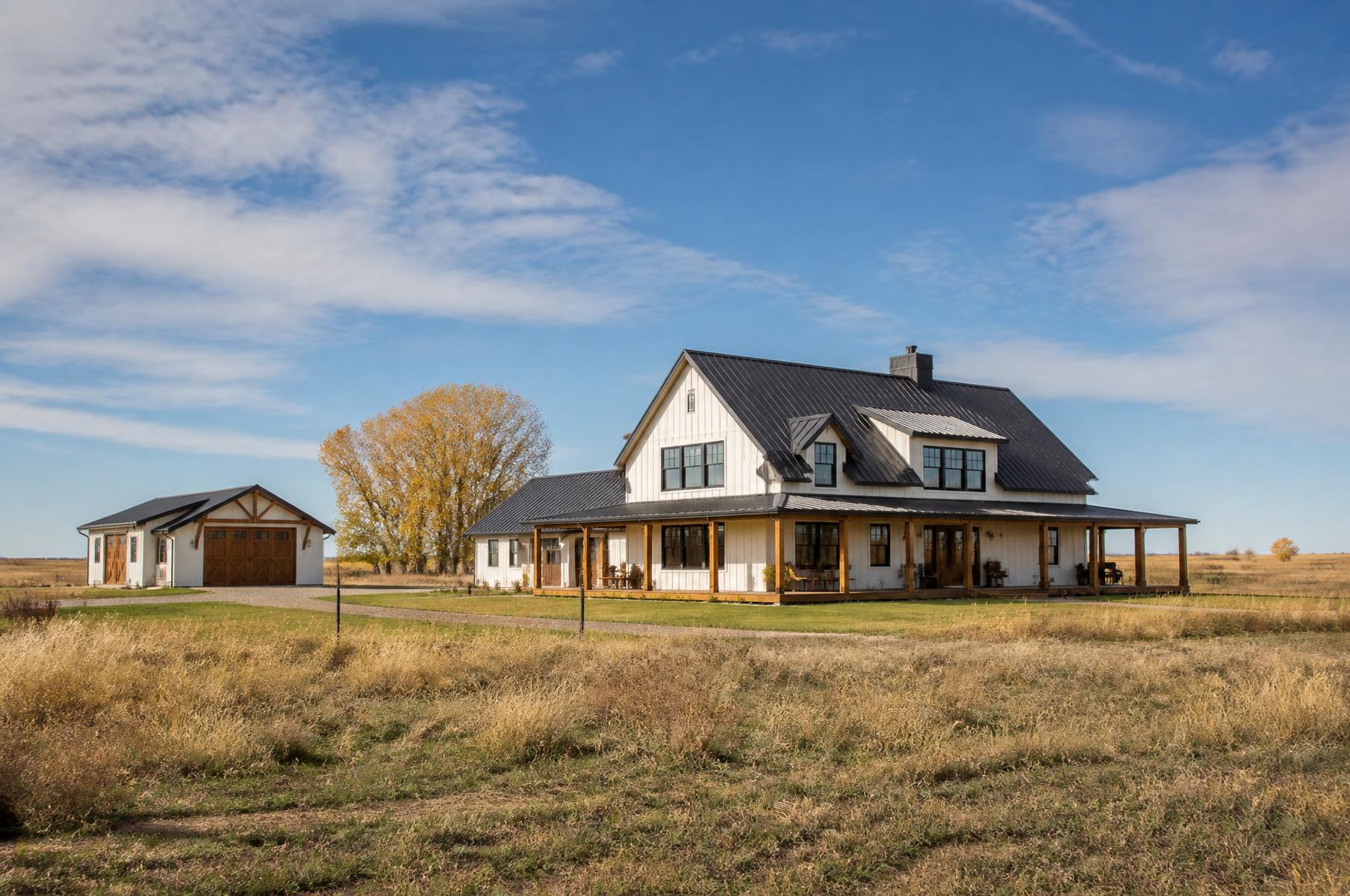 Completed white modern farmhouse with a wraparound porch and detached shop on open Colorado plains in fall