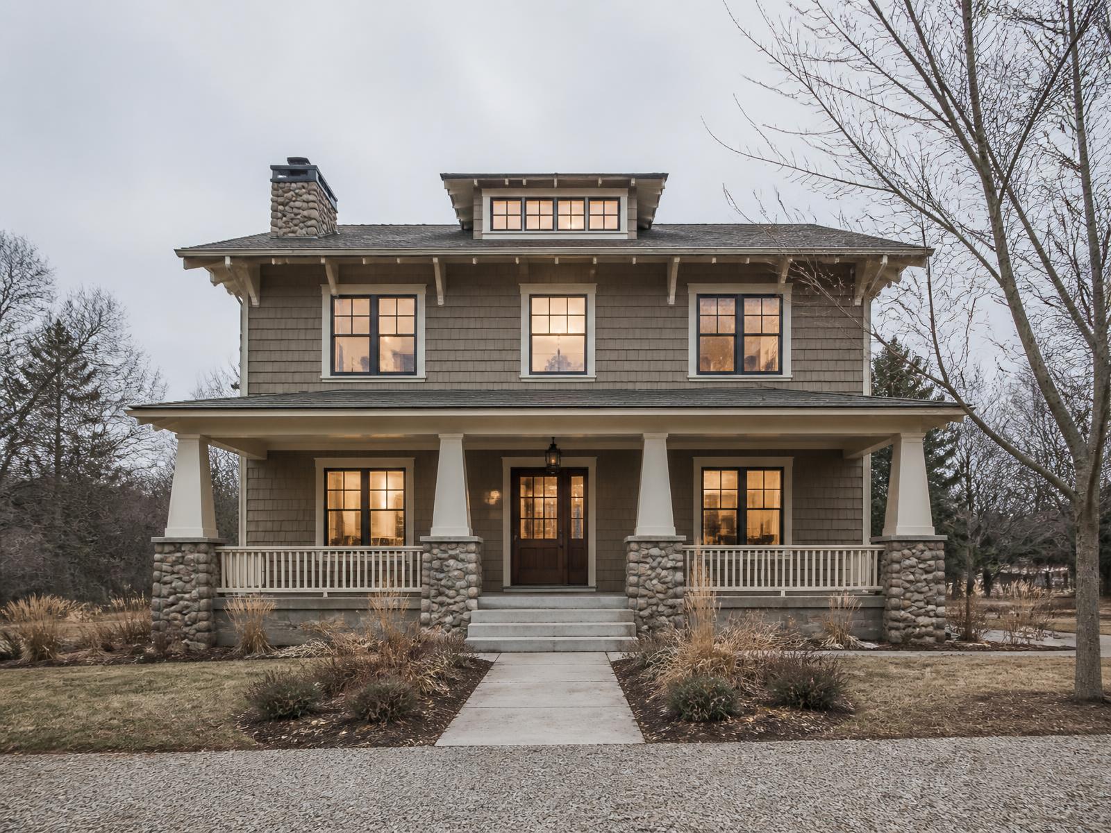 Two-story craftsman home concept with cedar shingle siding, cream trim, a shed dormer and a full-width porch on stone piers under an overcast sky
