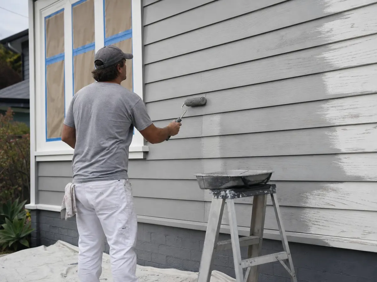 Exterior painter rolling grey paint onto lap siding next to a window masked with painter's tape